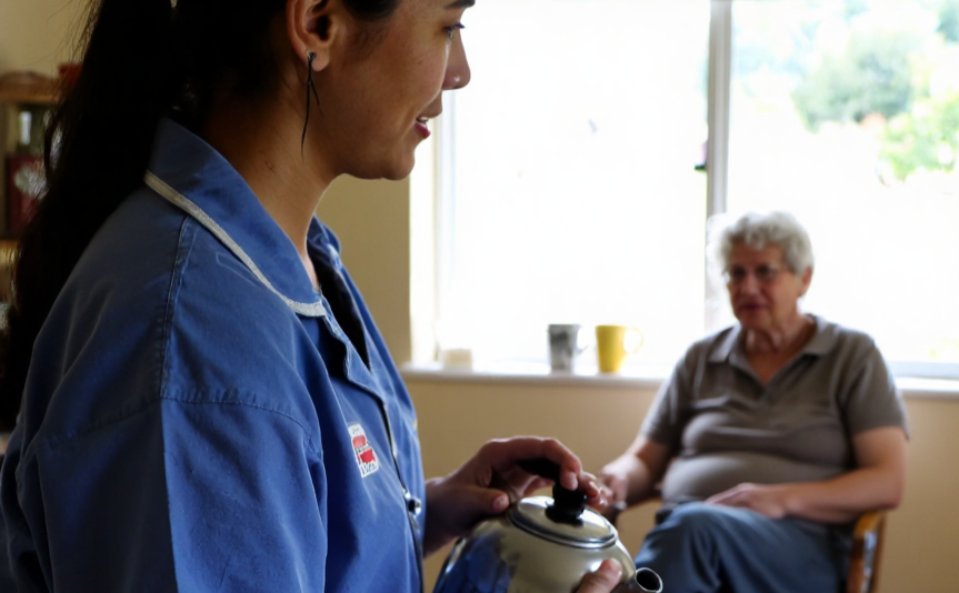 Younger New Zealander carer in a plain blue uniform putting a kettle on in a cosy kitchen while Mum sits relaxed at the table – in-home care in Christchurch and Tauranga