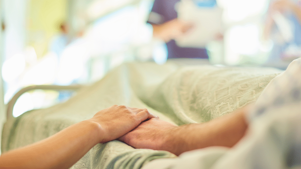 Close-up of a caregiver gently holding an older patient’s hand in a hospital bed, symbolising recovery and support after discharge in Christchurch and Tauranga