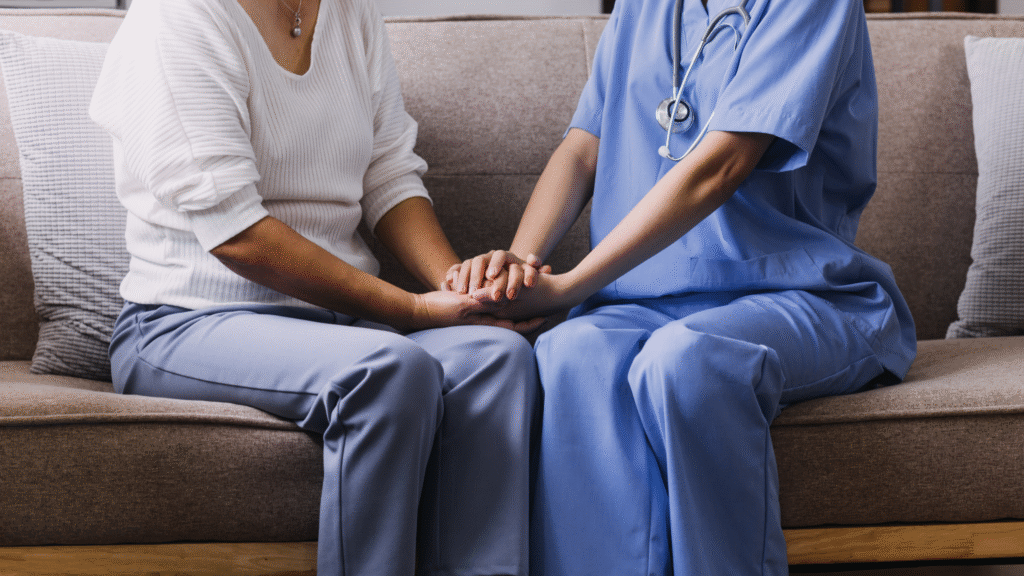 A New Zealand carer in a blue uniform holding hands with an older woman at home during a wellness check in Christchurch and Tauranga